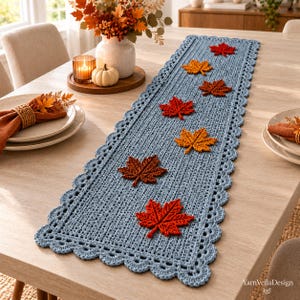 Puede incluir: Un camino de mesa rectangular de ganchillo azul claro con un borde festoneado y seis aplicaciones tridimensionales de hojas de arce en tonos rojos y naranjas dispuestas en línea por el centro. El texto YarnVellaDesign aparece en la esquina.