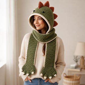 May include: A crochet hooded scarf in olive green with a cream-coloured trim. The hood features dark orange spikes along the top and back, and two black eyes. The scarf ends are shaped like dinosaur paws with cream-coloured claws at the tips.