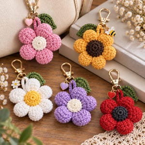 May include: Five crochet flower keychains with gold-tone lobster clasps and green leaves. The flowers are pink, yellow, white, purple, and red, each featuring a contrasting centre and a small charm, including hearts, a daisy, and a bee.