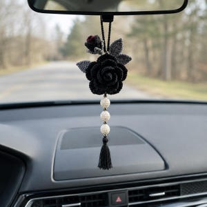 May include: A hanging car ornament featuring a large black crocheted rose with a smaller dark red bud and grey leaves. Below the flower are three cream-coloured textured beads and a black thread tassel, all suspended from a black cord.