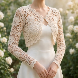 May include: A cream-colored, long-sleeved crochet bolero jacket featuring an intricate floral pattern throughout the knit. The garment has a scalloped edge along the front opening and is fastened at the neckline with a single, small, round white pearl button.