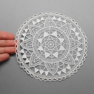 Peut inclure: Une dentelle de papier blanche avec un motif délicat et complexe. La dentelle est circulaire et présente un motif central en étoile entouré de motifs plus petits et répétitifs.