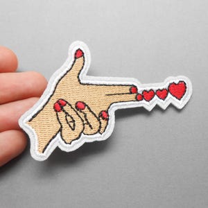 Peut inclure: Écusson brodé représentant une main en forme de pistolet, avec du vernis à ongles rouge et une rangée de cœurs rouges. L'écusson a une bordure blanche et est sur fond gris. Pour vêtements ou accessoires.