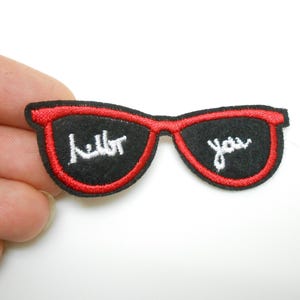 Peut inclure: Patch thermocollant brodé rouge et noir de lunettes de soleil avec les mots "Hello You" en blanc.