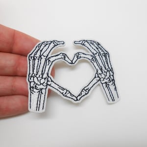 Peut inclure: Écusson brodé représentant un cœur formé par deux mains de squelette. Le patch a un fond blanc avec des détails noirs, créant un thème gothique ou d'Halloween. Idéal pour les vêtements ou accessoires.