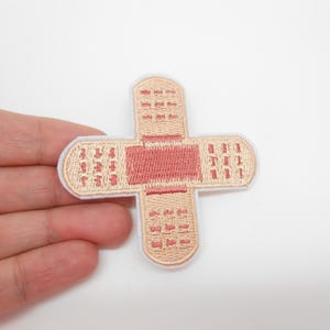 Peut inclure: Écusson brodé en forme de croix, ressemblant à un pansement. Le patch présente un fond beige clair avec un centre et des détails roses. Les bords sont soulignés d'une couleur beige plus claire.