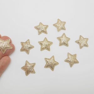 Peut inclure: Plusieurs embellissements en forme d'étoiles dorées à paillettes. Les étoiles sont dispersées sur une surface blanche, avec une étoile tenue dans une main. Les étoiles ont un aspect texturé et scintillant, parfait pour les travaux manuels et les décorations.