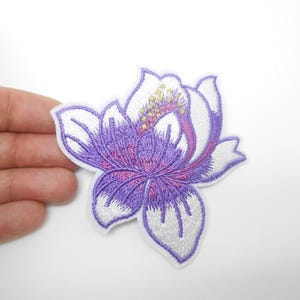 Peut inclure: Écusson floral brodé représentant une fleur violette et blanche avec des détails complexes. La fleur a un contour blanc et un centre violet avec des accents roses et jaunes. Idéal pour décorer des vêtements ou des accessoires.