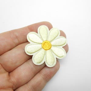 Peut inclure: Écusson brodé en forme de marguerite avec un centre jaune et des pétales blanc cassé. L'écusson est tenu dans une main sur fond blanc. Ce patch floral peut être utilisé pour décorer des vêtements, des sacs ou d'autres articles en tissu.