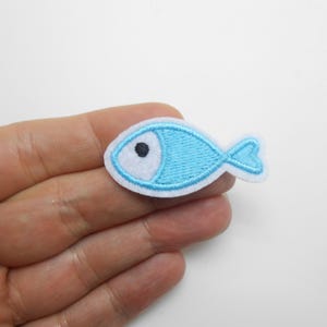 Peut inclure: Un petit écusson brodé en forme de poisson. Le poisson est bleu clair avec un bord blanc et un œil noir. L'écusson est tenu dans une main sur fond blanc.