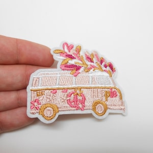 Peut inclure: Écusson brodé représentant un van rose et doré avec le mot "hippie". Le van est décoré de fleurs et d'un symbole de paix. Un bouquet de fleurs roses et dorées est posé sur le toit.