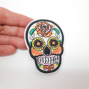 Peut inclure: Écusson brodé en forme de crâne avec un contour noir, présentant un motif coloré. Le crâne a des accents orange, jaunes et roses, avec une rose sur le dessus. L'écusson est sur fond blanc.