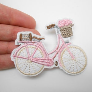 Peut inclure: Patch brodé de couleur rose représentant un vélo avec un panier brun rempli de fleurs roses. Le vélo a des roues blanches, une selle et un guidon bruns.