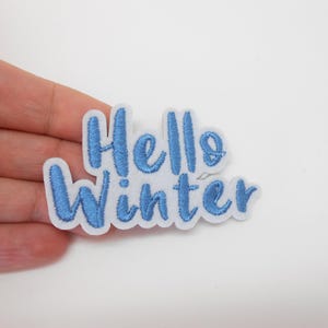 Peut inclure: Un écusson en feutre blanc brodé des mots "Hello Winter" en fil bleu clair. L'écusson a une bordure blanche. L'écusson est tenu dans la main d'une personne.