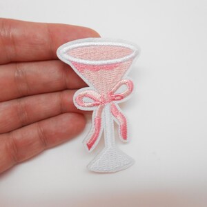 Peut inclure: Écusson brodé représentant un verre à cocktail rose avec un bord et une base blancs. Un nœud rose orne la tige. Le patch a une bordure blanche et est probablement destiné à la décoration.