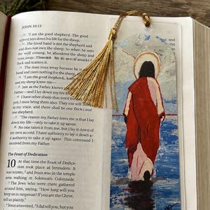 May include: A clear plastic bookmark featuring a painted illustration of a figure with a halo wearing a red robe, walking through blue water. The bookmark is topped with a gold-colored tassel and is placed inside an open book with printed text.