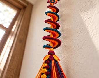May include: A hanging crochet decoration featuring a central spiral structure in orange, teal, and burgundy yarn. The top is finished with a small flower, and the bottom consists of a dense tassel with long yarn strands and three small yellow spheres.