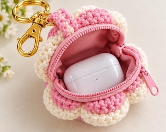May include: A flower-shaped crochet pouch in alternating cream and pink yarn, featuring a gold-tone metal lobster clasp keychain. The pouch is open, revealing a smooth pink fabric lining and a white rectangular wireless earbud charging case inside.
