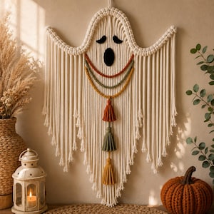 May include: A cream-colored macrame wall hanging shaped like a ghost, featuring black felt eyes and a mouth. It includes three curved braided ropes in rust, olive green, and mustard yellow, with three matching tassels hanging vertically below.