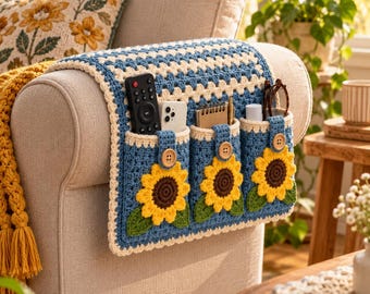 May include: A blue and cream crochet armchair caddy featuring three pockets, each decorated with a yellow and brown sunflower motif and a wooden button. The caddy is draped over the arm of a beige sofa and holds a remote, phone, and notebook.