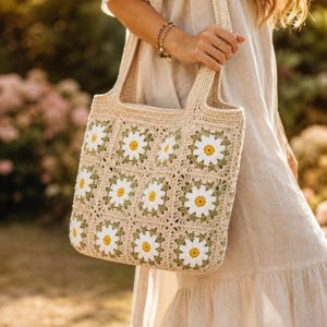 May include: A beige crochet tote bag featuring a repeating pattern of square motifs. Each square contains a white daisy with a yellow center on a sage green background. The bag has two integrated shoulder straps and a textured, handmade appearance.