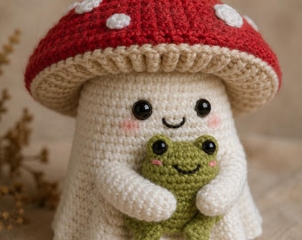 May include: A crocheted mushroom figure with a red cap featuring white spots and a cream-colored stem. The mushroom has a smiling face with black eyes and pink cheeks, and it is holding a small green crocheted frog with black eyes and pink cheeks.