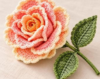 May include: A handcrafted crochet rose featuring layered petals in shades of orange, pink, and pale yellow with scalloped edges. The flower is attached to a green stem with two textured, pointed green leaves extending from the side.