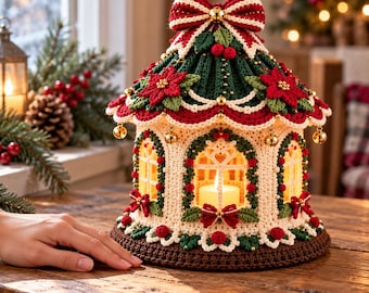 Könnte beinhalten: Eine gehäkelte Laterne mit cremefarbener Basis, dunkelgrünem Dach und roten Weihnachtsstern-Akzenten. Die Struktur zeigt Bogenfenster, kleine goldene Glöckchen am gewellten Rand und eine Schleife mit zentraler Goldglocke.