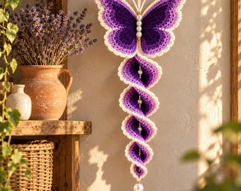 May include: A hanging crochet decoration featuring a purple butterfly at the top with white-edged wings and antennae. Below the butterfly, a spiraling, multi-layered purple crochet tail descends, accented with white pearls along the center spine.