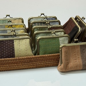 May include: A collection of rectangular coin purses with bronze-toned metal kiss-lock frames. The purses feature various fabric patterns including polka dots, small crosses, and solid textures in shades of brown, green, tan, and grey, arranged in a woven basket.