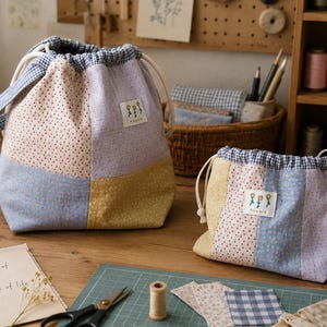 May include: Two drawstring pouches made of patchwork fabric in lavender, yellow, blue, and floral patterns. Each bag features a blue and white gingham trim at the top, white cord ties, and a small rectangular label with the text flowers.