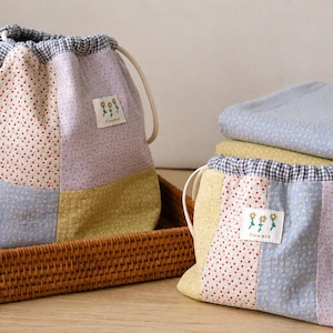 May include: Two drawstring pouches made of patchwork fabric in pastel shades of yellow, blue, and lavender with small floral patterns. Each pouch features a blue and white gingham trim and a white rectangular label with three flowers and the text "flowers".