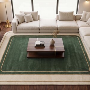 May include: A rectangular area rug featuring a deep green center panel bordered by a thick cream-colored frame and a thin gold-toned line. The rug has rounded corners and is positioned on a dark wood floor beneath a wooden coffee table.