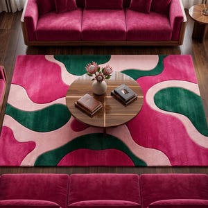 May include: A rectangular area rug featuring an abstract, fluid pattern of interlocking shapes in vibrant magenta, deep forest green, and soft pale pink. The rug is positioned centrally on a dark wood floor, surrounded by matching magenta velvet furniture.