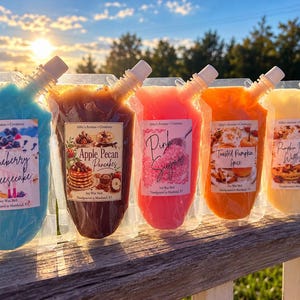 May include: Five clear plastic pouches filled with colorful soy wax melts, each featuring a label with the text Abby's Aromas + Creations and specific scent names: Blueberry Cheesecake, Apple Pecan Pancakes, Pink Sugar, Toasted Pumpkin Spice, and Pumpkin Pecan Waffles.