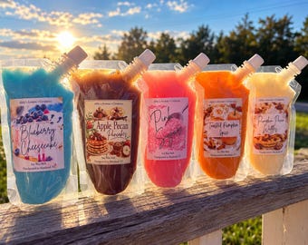 May include: Five clear plastic pouches filled with colorful soy wax melts, each featuring a label with the text Abby's Aromas + Creations and specific scent names: Blueberry Cheesecake, Apple Pecan Pancakes, Pink Sugar, Toasted Pumpkin Spice, and Pumpkin Pecan Waffles.