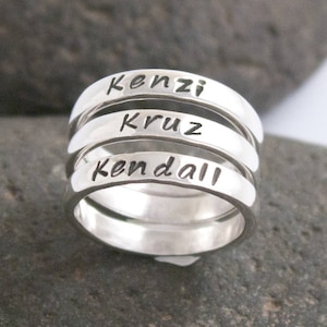 May include: Four silver rings stacked on top of each other. The rings are engraved with the names Kenzi, Cruz, and Kendall.
