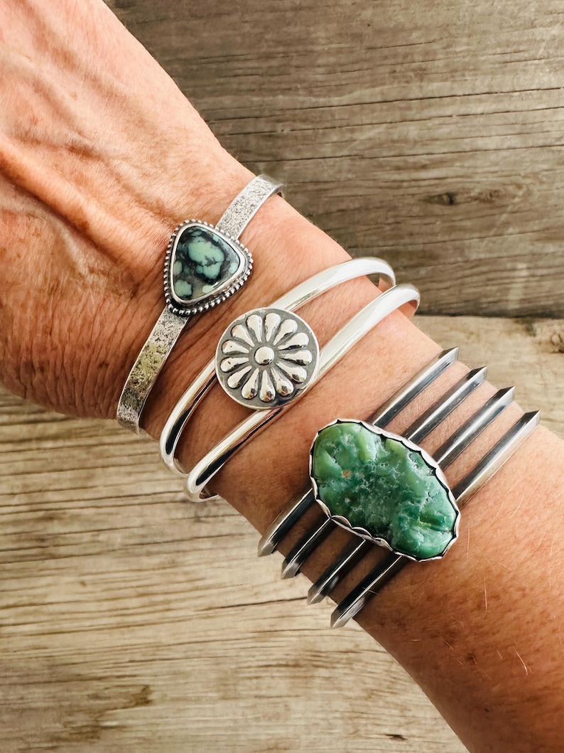 Puede incluir: Cuatro pulseras de plata con piedras turquesa. Las pulseras est&aacute;n apiladas en la mu&ntilde;eca de una persona. Las piedras turquesa son de diferentes formas y tama&ntilde;os. Las pulseras est&aacute;n hechas de plata de ley y tienen un aspecto r&uacute;stico.