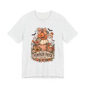 May include: A light gray t-shirt featuring a graphic of stacked pumpkins, a small ghost wearing a beanie, autumn flowers, and bats. The text reads THE PUMPKIN PATCH SOCIAL, surrounded by a decorative border with a plaid bow at the top and bottom.