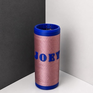 May include: A cylindrical beverage cooler featuring a metallic rose gold textured exterior and a solid blue rim at the top and bottom. The name JOEY is displayed in bold, raised blue capital letters across the center of the rose gold surface.