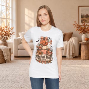 May include: A white short-sleeved t-shirt featuring a colourful graphic of stacked pumpkins, a small ghost, sunflowers, and autumn foliage. The text "THE PUMPKIN PATCH SOCIAL" is printed in a decorative font at the base of the illustration.