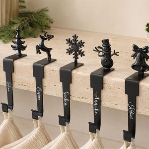 May include: Five matte black metal stocking holders with personalized names engraved on the vertical stems. Each holder features a unique holiday-themed topper: a Christmas tree, a reindeer, a snowflake, a snowman, and a bell, all hanging from a ledge.
