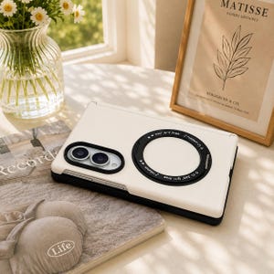 Puede incluir: Una funda plegable para smartphone de color blanco con un módulo de cámara negro y un anillo magnético circular negro en la parte posterior. El anillo tiene impreso el texto 360 degree rotation e iconos de notas musicales. Borde negro.