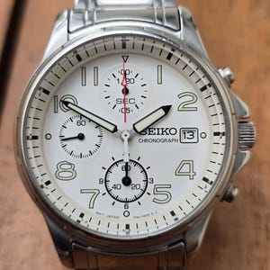 May include: A silver-tone Seiko chronograph watch with a white dial, featuring black Arabic numerals, three sub-dials, a date window at the three o'clock position, and a stainless steel link bracelet. The watch face displays the text Seiko Chronograph.