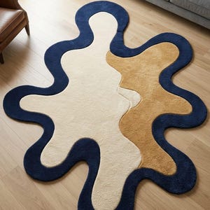 May include: An area rug with an irregular, organic shape featuring a thick navy blue border. The interior of the rug is divided into two distinct sections, one in a creamy off-white color and the other in a warm tan, both with a plush texture.