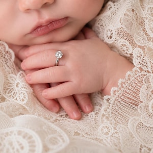 May include: A silver-coloured ring featuring a single round-cut clear gemstone set in a prong setting. The ring is positioned on a small finger, resting against a delicate, cream-coloured lace fabric with intricate floral patterns and scalloped edges.