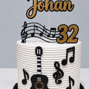 May include: A white cake with horizontal ridges, decorated with black and gold musical notes, a guitar cutout, and a custom topper featuring the name Johan and the number 32 in black and gold script and block lettering on a thin stick.