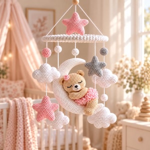 May include: A crochet baby mobile featuring a central white crescent moon with a sleeping tan bear wearing a pink tutu and flower. The mobile includes hanging white clouds, pink and gray stars, and small white and pink spheres suspended from a ring.