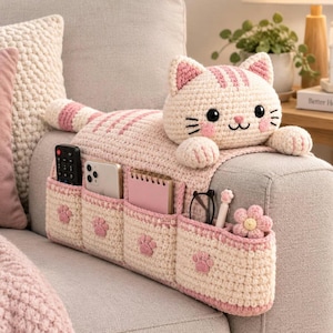 May include: A cream and light pink crocheted cat-shaped sofa armrest organizer featuring four storage pockets. Each pocket is decorated with a small pink paw print applique. The cat head has black eyes, a pink nose, and pink striped ears.