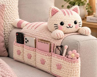 May include: A cream and light pink crocheted cat-shaped sofa armrest organizer featuring four storage pockets. Each pocket is decorated with a small pink paw print applique. The cat head has black eyes, a pink nose, and pink striped ears.